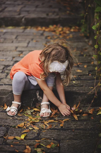 Linda niña en cárdigan naranja jugando y recogiendo hojas mientras está sentada en un viejo camino de piedra con escaleras — Foto de Stock