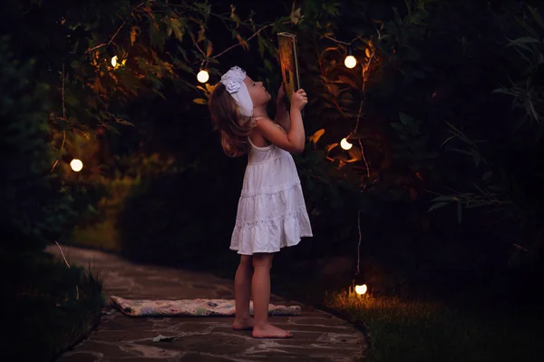 Чарівна дівчинка в білій сукні тримає книгу в вечірньому літньому саду, прикрашеному вогнями — стокове фото
