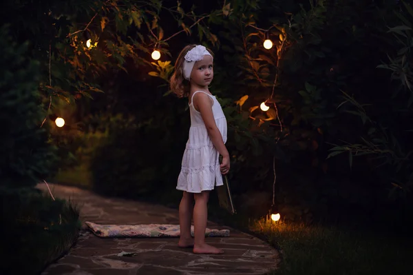 Adorabile bambina in abito bianco libro di lettura nel giardino estivo serale decorato con luci — Foto Stock