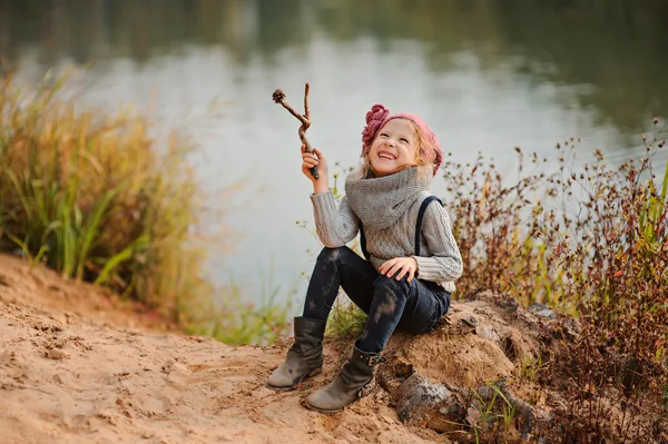 Очаровательная улыбающаяся девочка в розовой вязаной шляпе сидит с палкой на берегу реки с песчаным пляжем — стоковое фото