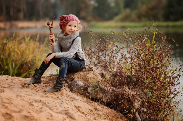 Adorable smiling child girl in pink knitted hat sitting with stick on river side with sand beach — Stock Photo, Image