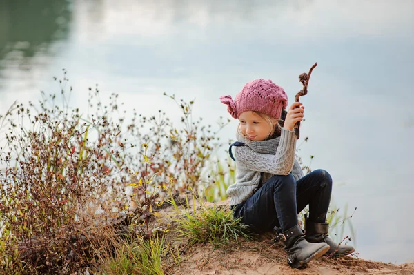 Bedårande leende barn flicka i rosa Stickad mössa sitter med pinne på flodsidan med sandstrand — Stockfoto