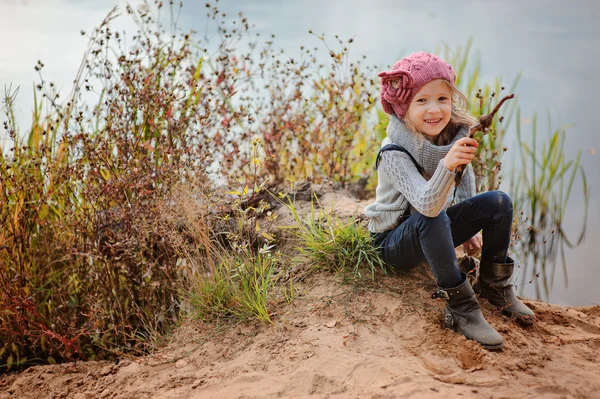 Adorable niña sonriente en rosa sombrero de punto sentado con palo en el lado del río con playa de arena — Foto de Stock