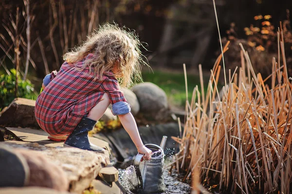 Child girl in plaid dress gathering water from pond in spring garden — Stock Photo, Image