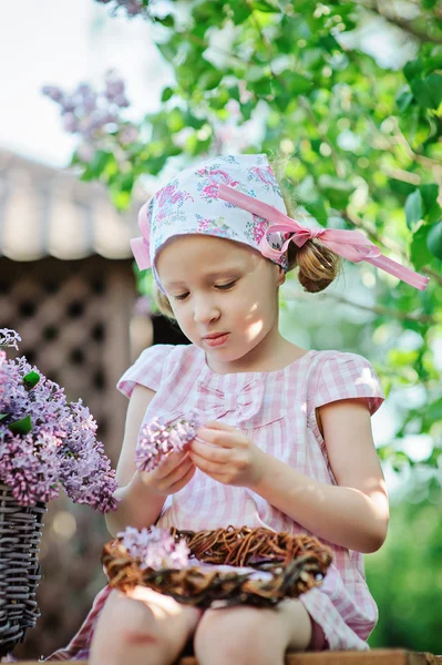 Adorable niña en vestido de cuadros de color rosa haciendo corona de color lila en el jardín floreciente primavera — Foto de Stock