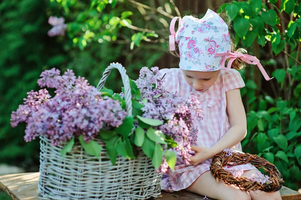 Adorable child girl in pink plaid dress making lilac wreath in spring blooming garden — Stock Photo, Image