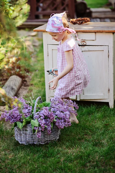 Menina bonito no jardim da primavera com cesta de lilases perto de bureau de madeira vintage — Fotografia de Stock