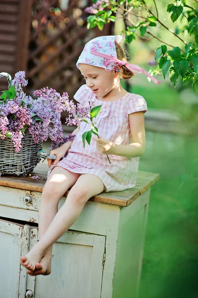 Spring portrait of adorable child girl in pink plaid dress cuts lilacs in sunny garden — Stock Photo, Image
