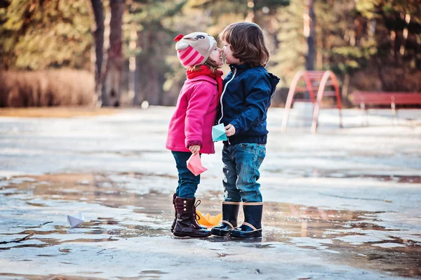 Toddler boy and girl having fun and kisses holding paper boats in spring puddle — Stock Photo, Image