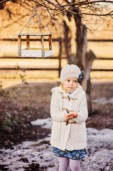 Vertical outdoor portrait of adorable blonde child girl in beige knitted coat and hat with bird feeder — Stock Photo, Image