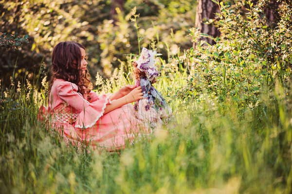 Dreamy kid girl dressed as fairytale princess playing with her doll in summer forest