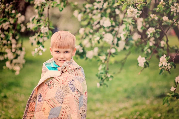 Glücklicher Junge in kuschelige Decke gehüllt genießt den Frühling im blühenden Garten — Stockfoto