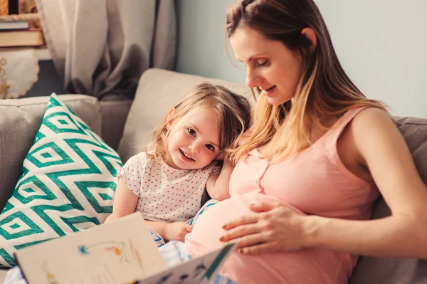 Glücklich liebende schwangere Mutter liest ihrer kleinen Tochter zu Hause auf dem Sofa vor — Stockfoto