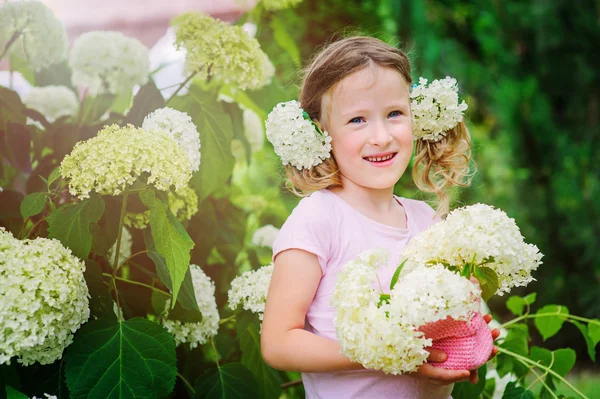 Cute happy child girl playing with hydrangea flowers in pink knitted basket in summer garden near flowering bush — Stock Photo, Image