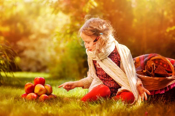 Fröhliches Kindermädchen in weißem Strickschal sitzt mit roten Äpfeln im sonnigen Herbstgarten — Stockfoto