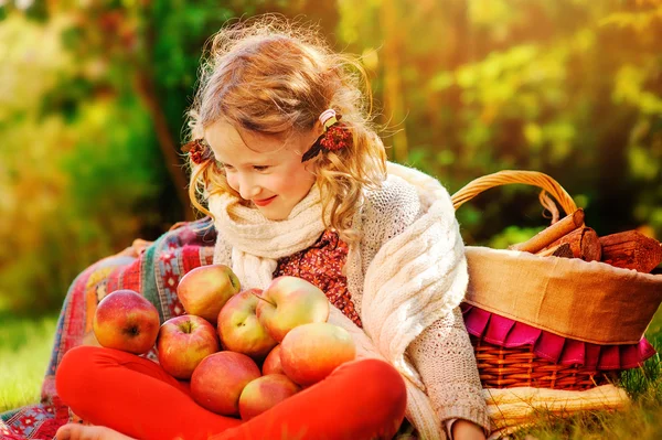 Happy child girl in white knitted scarf sitting with red apples in sunny autumn garden — Stock Photo, Image