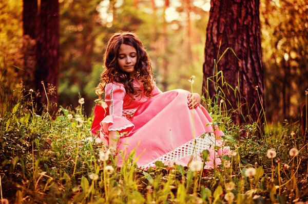 Curly child girl in pink fairytale princess dress sitting in summer forest