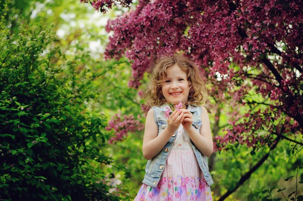 Gelukkig kind meisje in jurk spelen in de buurt van bloeiende boom in voorjaar tuin. Rural gezellige scène, seizoensgebonden activiteiten in de buitenlucht. — Stockfoto