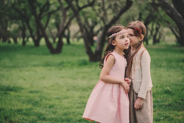 two kid girlfriends telling secrets to each other in spring park. Little dressy sisters spending time outdoor