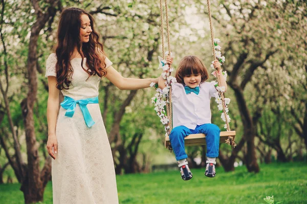 Mãe amorosa com filho criança feliz no balanço no jardim de primavera ensolarado. Família a passar tempo juntos. Atividades ao ar livre sazonais . — Fotografia de Stock