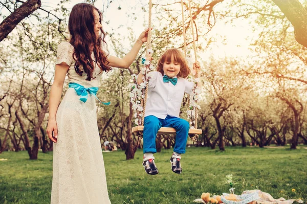 Liebevolle Mutter mit glücklichem Kleinkind-Sohn auf Schaukel im sonnigen Frühlingsgarten. Familie verbringt Zeit miteinander. Saisonale Aktivitäten im Freien. — Stockfoto