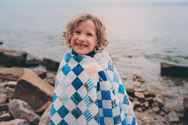 Linda niña rizada feliz relajarse en la playa de piedra, envuelto en una acogedora manta de edredón. Actividades de vacaciones de verano, viajar en Europa de vacaciones . — Foto de Stock