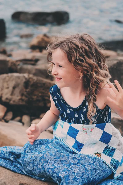 Linda niña rizada feliz relajarse en la playa de piedra, envuelto en una acogedora manta de edredón. Actividades de vacaciones de verano, viajar en Europa de vacaciones . — Foto de Stock