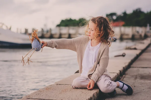 Linda niña rizada relajándose en la playa y jugando con el pájaro de juguete. Viajar a Portoroz, Eslovenia en vacaciones de verano . — Foto de Stock