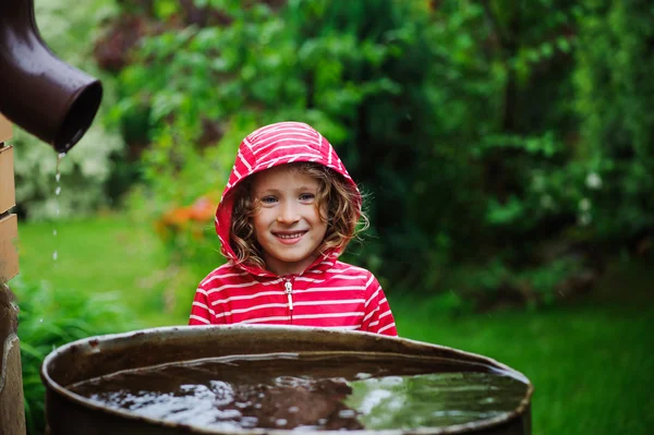 Niña en impermeable rojo jugando con el barril de agua en el jardín de verano lluvioso. Economía del agua y cuidado de la naturaleza — Foto de Stock