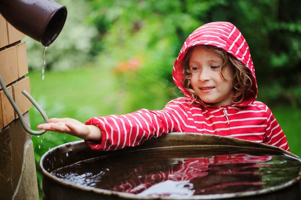Çocuk kız kırmızı yağmurluk ile su varil yağmurlu yaz bahçesinde oynarken. Su Ekonomi ve doğa bakım kavramı — Stok fotoğraf