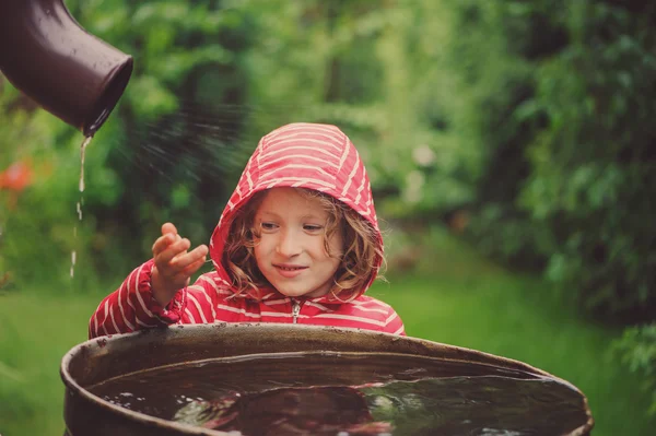 雨の夏の庭での水のバレルで遊んで赤いレインコートの子供女の子。水経済と自然ケアのコンセプト — ストック写真