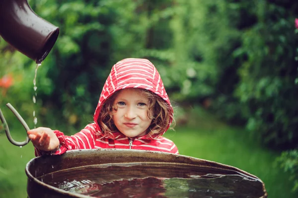 Enfant fille en imperméable rouge jouant avec baril d'eau dans le jardin d'été pluvieux. Economie de l'eau et concept de protection de la nature — Photo