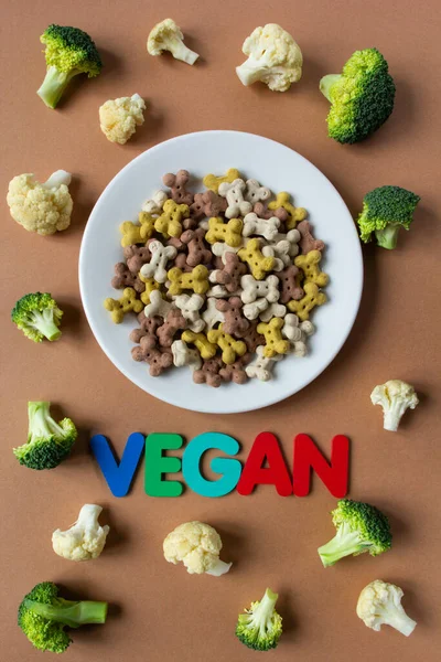Hond vegetarische droge crunchies op plaat en groenten op beige achtergrond met belettering VEGAN. — Stockfoto