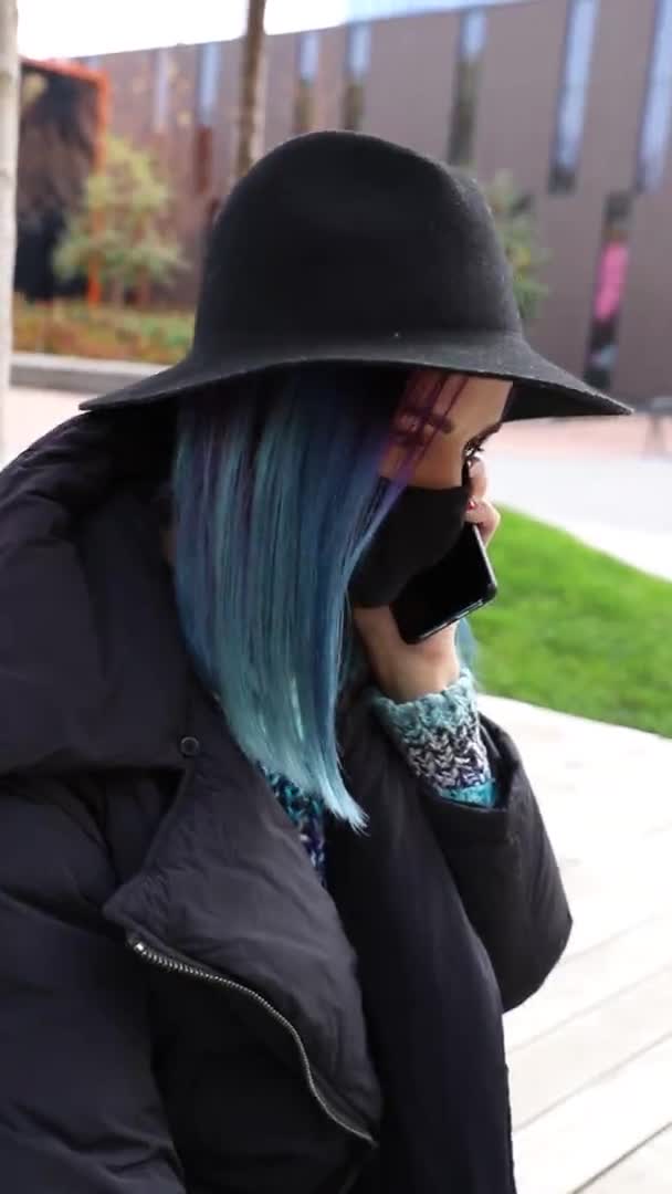 Μαθήτρια με μπλε μαλλιά σε προστατευτική μαύρη μάσκα και καπέλο που μιλάει στο τηλέφωνο έξω. Κάθετη βίντεο — Αρχείο Βίντεο