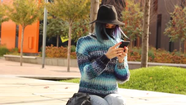 Девушка с голубыми волосами в защитной черной маске и шляпе со смартфоном. Городской модный костюм. — стоковое видео