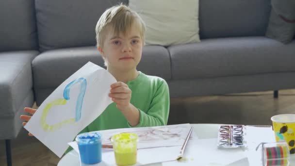 Down sendromlu çocuk elinde sarı ve mavi kalpli bir resim tutuyor. — Stok video
