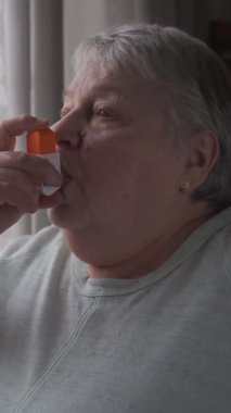 Yaşlı kadın evde astım spreyi kullanıyor. Dikey biçim