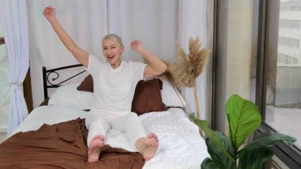 Emocional feliz mujer mayor sentada en la cama, celebra las buenas noticias, se divierten — Vídeos de Stock