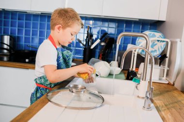 Önlüklü bir çocuk evde bulaşık yıkıyor.
