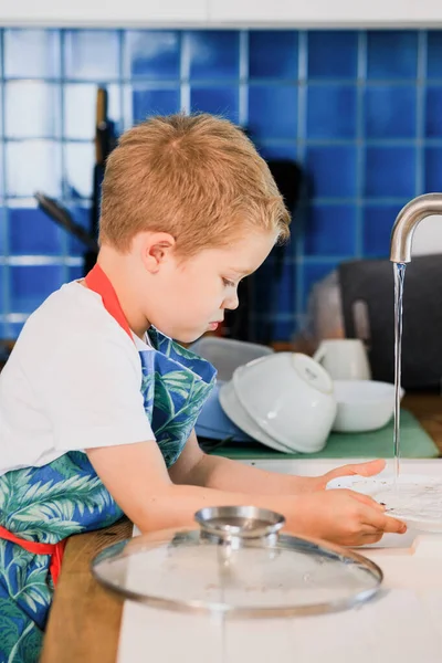 Egy kötényes fiú mosogatott otthon a konyhában. — Stock Fotó