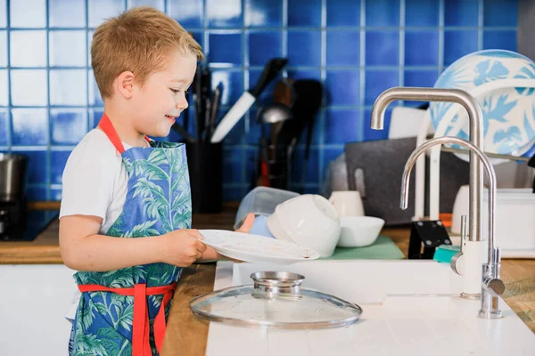 Un niño en un delantal lava los platos en la cocina en casa Fotos De Stock Sin Royalties Gratis