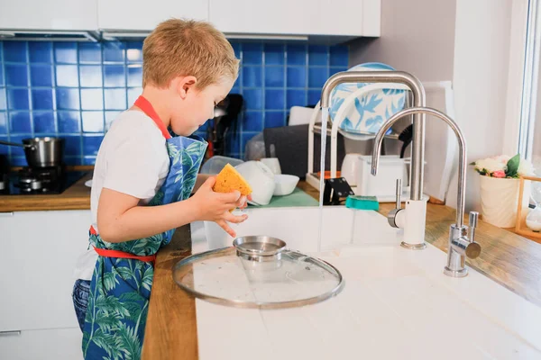 Un niño en un delantal lava los platos en la cocina en casa Imagen De Stock