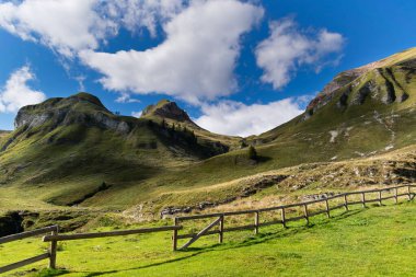 Etkileyici dağ manzarası, beyaz bulutlu mavi gökyüzü, çobanın kulübesini çevreleyen ahşap çitli yeşil yamaçlar. Piani Eterni, Dolomiti Bellunesi Ulusal Parkı, İtalya