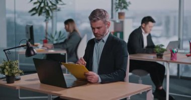 Beyaz Sakallı Yetişkin İşadamı 'nın Laptop Kullanan ve Kameraya Gülümseyen Meslektaşlarla İşbirliği Yapan Notları Yazan Portresi. Başarı ve İnsanlar.