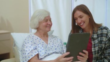 Yaşlı beyaz kadın hasta ve kızı dijital tabletle sohbet ediyorlar. Hastaneden akrabalarıyla konuşuyorlar. Aile desteği. Çevrimiçi iletişim.