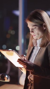 Kendini işine adamış bir iş kadını, geceleri modern bir ofis ortamında şık tabletindeki önemli verileri dikkatle analiz ediyor.