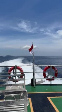 Feribot pruvası Okyanus Uyanışı, Günışığı Güvertesi ve Metal korkuluklu, İnce Bulutlara Karşı Çırpınan Kızıl Bayrak, Ufuk 'taki Uzak Ada, Derin Mavi Su, Hafif Sprey ve Serin Yaz Atmosferi