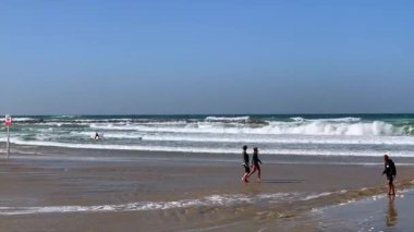 Güneşli sörfler kumsalda yürüyordu. Hafif dalgalar kıyıları süpürüyordu. Mavi gökyüzü, yansıtıcı kumlar, uzak cankurtaran bayrağı, günlük plaj giysisi, rahat adımlar, ritmik sörf sesleri, açık sahil havası.