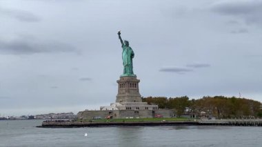 Limanda yükselen Özgürlük Anıtı, karamsar bulutlu gökyüzü, sakin dalgalı su, ön plandan geçen feribot, sonbahar ağaçlı kaide ve ada, ufukta Manhattan ufuk çizgisi, teknelerde ve sahilde ziyaretçiler,