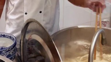 Asyalı şef zengin et suyunu kaynatıyor ve tezgahta erişte tabakları kaplıyor, porselen kaselere kepçe karıştırıyor, soğan ve kişniş malzemeler ekliyor, buhar yükseliyor, odaklanmış eller hızla hareket ediyor.,